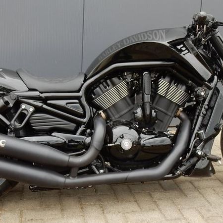 Ein schwarzes Custom-Motorrad mit matter und glänzender Oberfläche, geparkt auf einer gepflasterten Fläche vor einer grauen Wand.