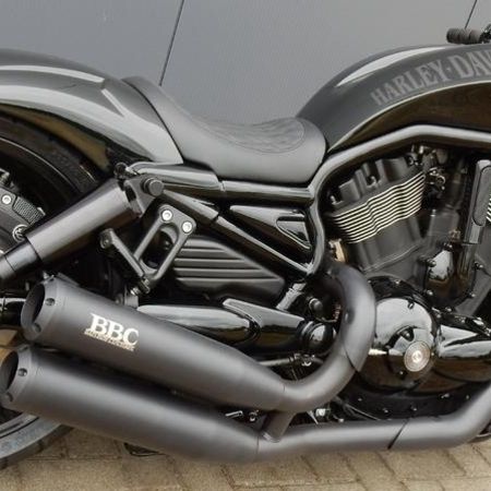 Ein schwarzes Custom-Motorrad mit matter und glänzender Oberfläche, geparkt auf einer gepflasterten Fläche vor einer grauen Wand.