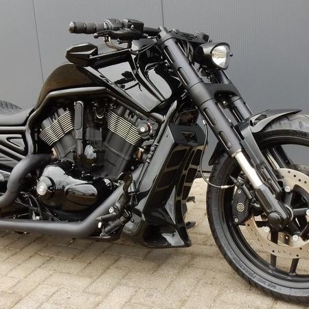 Ein schwarzes Custom-Motorrad mit matter und glänzender Oberfläche, geparkt auf einer gepflasterten Fläche vor einer grauen Wand.