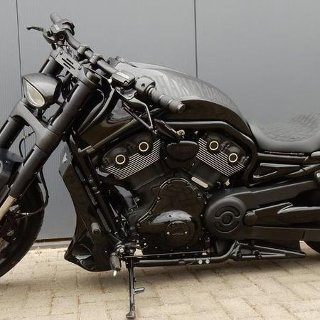 Ein schwarzes Custom-Motorrad mit matter und glänzender Oberfläche, geparkt auf einer gepflasterten Fläche vor einer grauen Wand.
