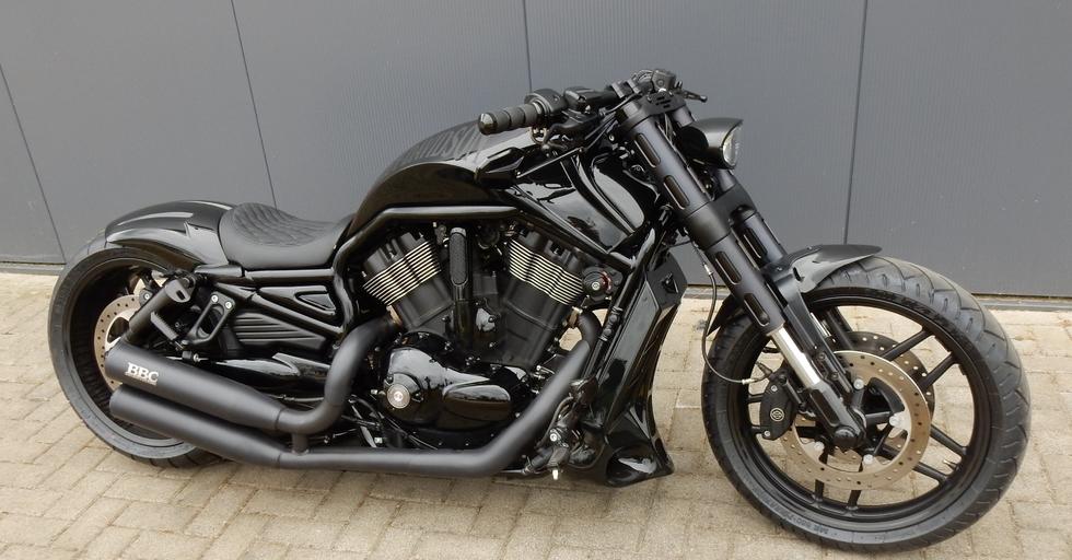 Ein schwarzes Custom-Motorrad mit matter und glänzender Oberfläche, geparkt auf einer gepflasterten Fläche vor einer grauen Wand.