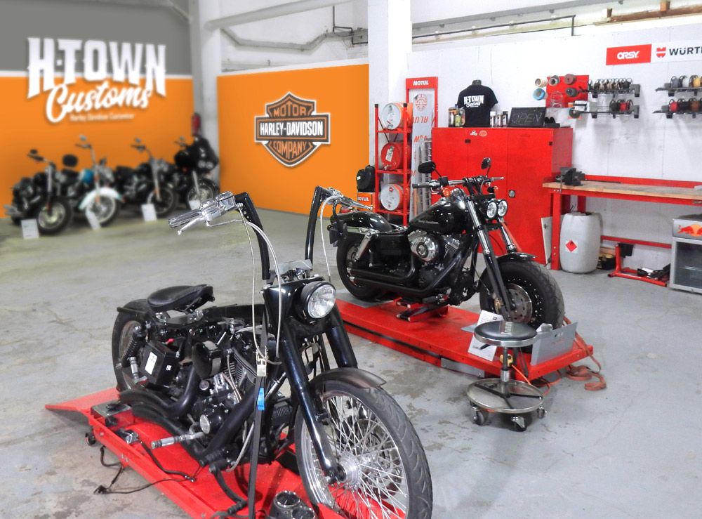 Zwei Custom-Motorräder auf roten Hebebühnen in einer Garage, mit einem Harley-Davidson-Schild im Hintergrund und weiteren Motorrädern in Sichtweite.